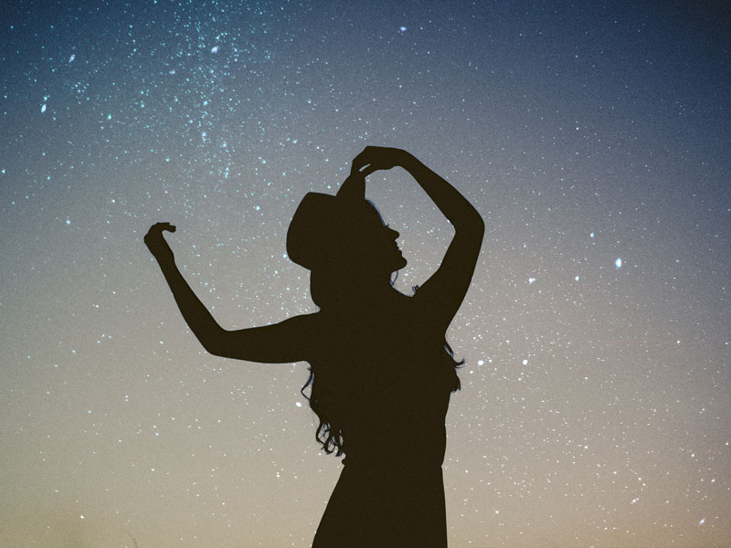 Silhouette of a person wearing a hat, with one hand raised, set against a starry night sky background. The individual appears to be posing gracefully, creating a contrast between their dark outline and the illuminated stars—an inspiring image for small business coaching sessions.