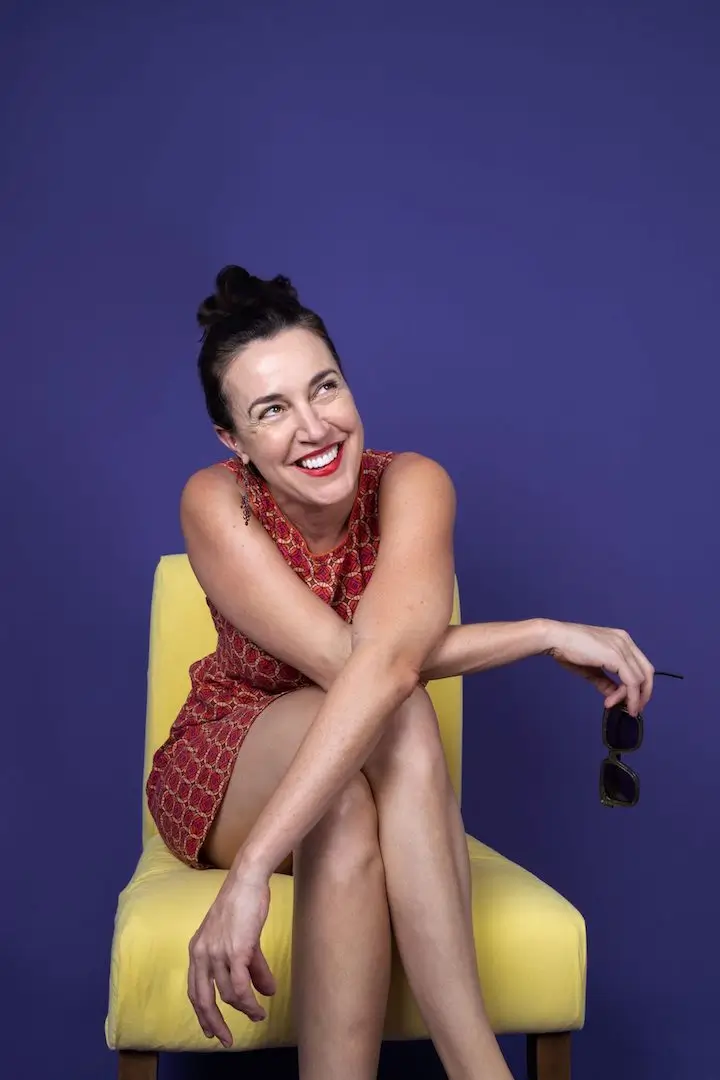 Brook McCarthy sits on a yellow chair in front of a purple background and wearing a red dress