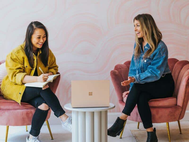 Two women sit on pink chairs and discuss something. They are smiling and one woman is writing in a notebook.